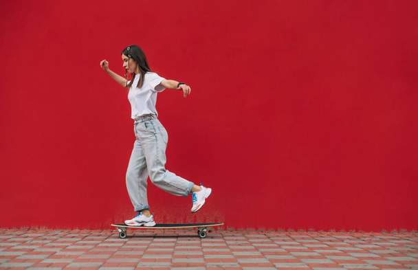 Woman skater in stylish street clothes skates on a background of red wall. Copy space - Fotografie, Obrázek