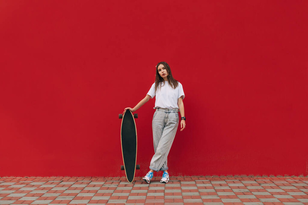 Portrait of an attractive female skater in stylish street clothes stands on a red background with a longboard in his hand and poses for the camera. Women's skateboarding. - Photo, Image