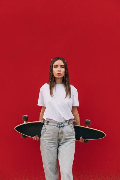 Portrait of stylish woman in street clothes isolated on red background with longboard in hands behind back, looking at camera with serious face. - Photo, Image