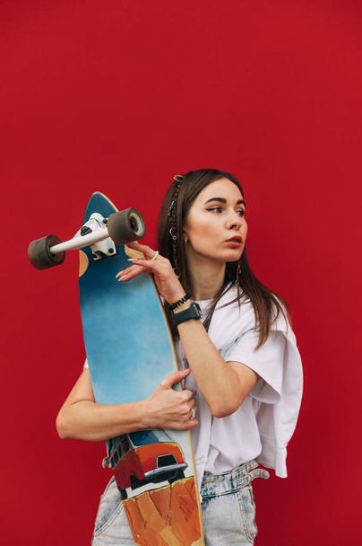 Portrait of stylish woman in casual clothes standing on red background with longboard in hands, looking aside with serious face and posing at camera. - Photo, Image