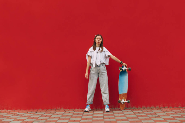 Woman in street clothes stands with a longboard in her hand on a red background and poses for the camera with a serious face. Women's skateboarding. - Photo, Image