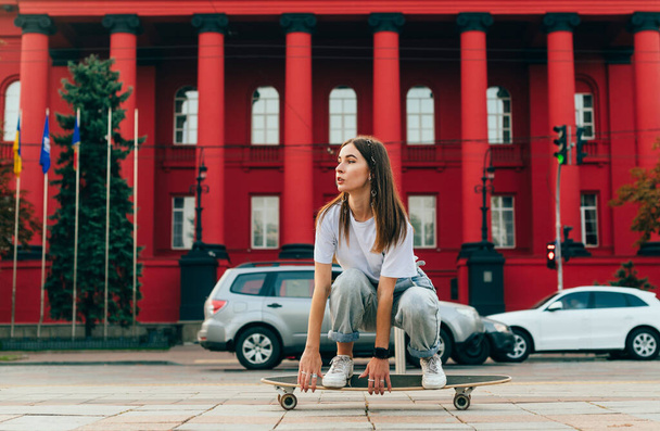 Piękna kobieta skater w casual ubrania na prędkości jazdy po ulicy na longboard i wygląda na boku. Kobieta łyżwiarka spacerująca po longboardzie na tle czerwonej architektury. - Zdjęcie, obraz