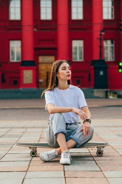 Portrait of an attractive stylish woman in casual clothes resting sitting on a longboard on the background of architecture, looking to the side with a serious face. - Foto, imagen