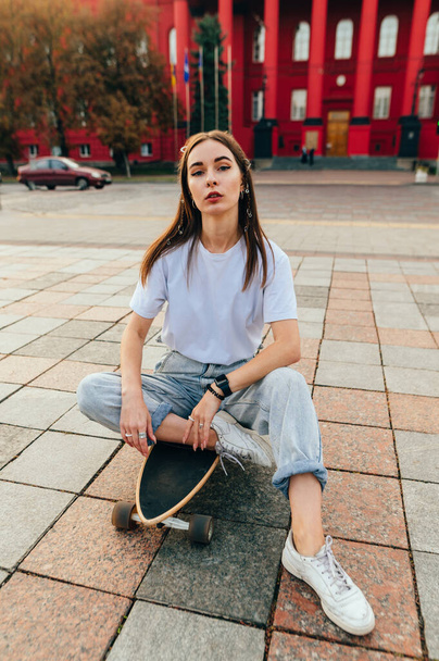 Stylish woman in casual clothes sits on a longboard outdoors after skiing, relaxing and looking at the camera with a serious face. - Photo, Image