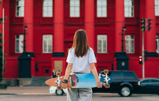 Costas de uma menina skatista elegante com um longboard atrás de suas costas fica em um fundo de arquitetura vermelha. Contexto - Foto, Imagem