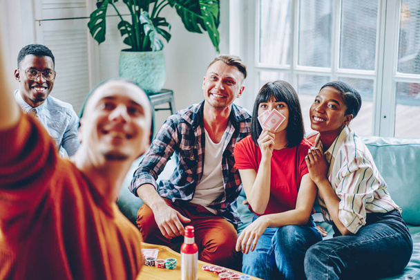 Happy multiracial friends posing for making selfie content during gather meeting for gambling and wagering with cards and poker chips, cheerful hipster guys enjoying together recreation in home flat - Fotó, kép