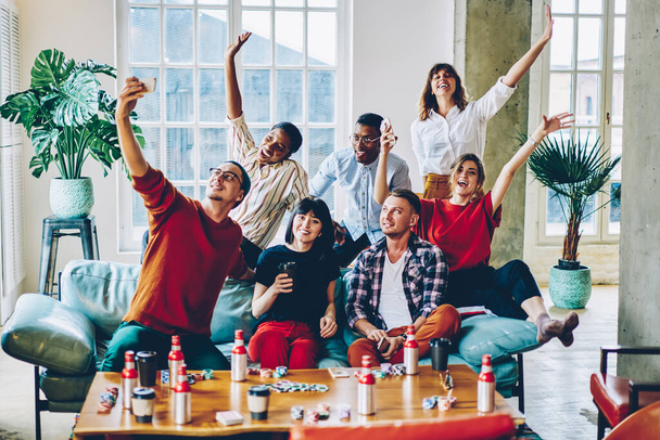 Happy multiracial friends in trendy clothing sitting at comfortable couch and raising hands while making selfie influence content, smiling poker players enjoying together leisure for wagering - Photo, image