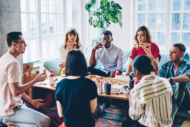 Hipster guys eating pizza spending free time together at home interior enjoying friendly company, young multicultural people dressed have party with Italian food and beer in modern apartment - Photo, Image