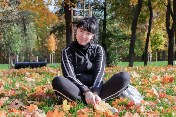 Negyvenedik évforduló csinos európai nő, tréningruhában, ül egy őszi parkban a füvön. - Fotó, kép