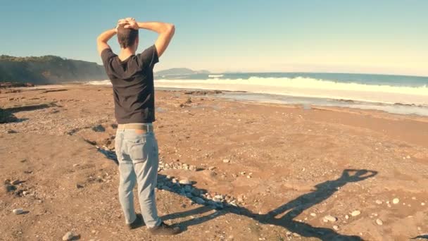 Typ in blauer Jeans und schwarzem Kurzarm-T-Shirt, der mit den Händen auf dem Kopf auf das Meer blickt - Filmmaterial, Video