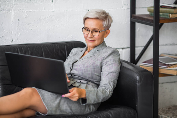 beautiful stylish adult woman in suit working with laptop on black sofa - Zdjęcie, obraz