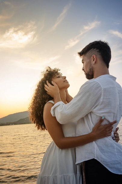 Young attractive caucasian bearded man with modern haircut holds his black Hispanic girlfriend's head in his hands looking into her eyes while she hugs him - Multiracial couple romance scene at sunset - Zdjęcie, obraz