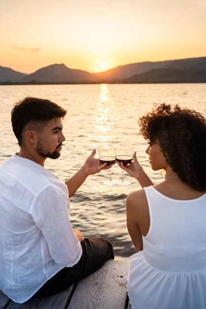 Romantische Szene eines gemischtrassigen Paares, das bei Sonnenuntergang auf einer Seebrücke sitzt oder im Morgengrauen mit Rotwein anstößt und sich gegenseitig in die Augen schaut - Attraktiver Mann, der sich mit seiner hispanischen Freundin zusammentut - Fokus auf Gläser - Foto, Bild