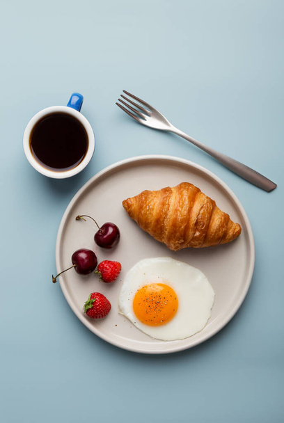 Breakfast with croissant, egg, coffee and berries at light blue minimal background. Concept of healthy tasty food. Top view, flat lay - 写真・画像