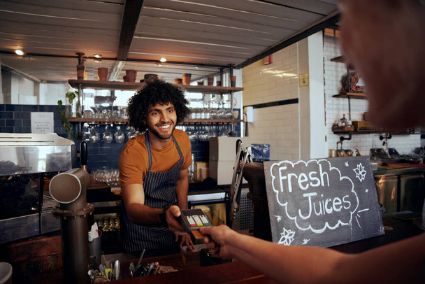 Portrait of smiling waiter holding credit card swipe machine while customer typing code in modern cafe - Zdjęcie, obraz