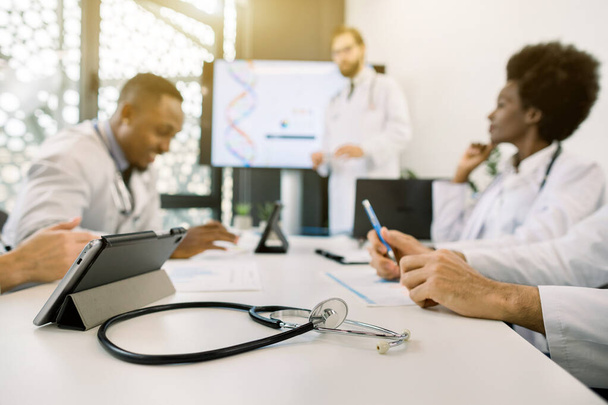 Medicine, technology concept. Blurred image of multiethnical male and female doctors having morning meeting in modern sunny office clinic. Focus on the stethoscope and digital tablet on the table - Foto, imagen