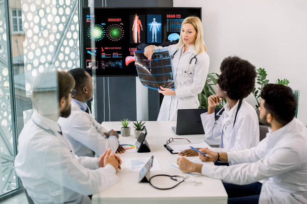 Pretty middle aged blond woman doctor has a speech at the hospital seminar, stands in front of digital screen with datas, holds CT and consults her multiethnic colleagues on diagnostics and treatment - Photo, Image