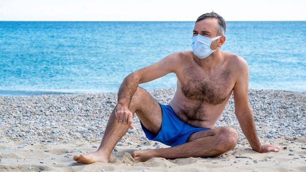 A man in white medical mask and blue swimming shorts with naked torso and chest hair sitting on a beach, sea on the background - Photo, Image