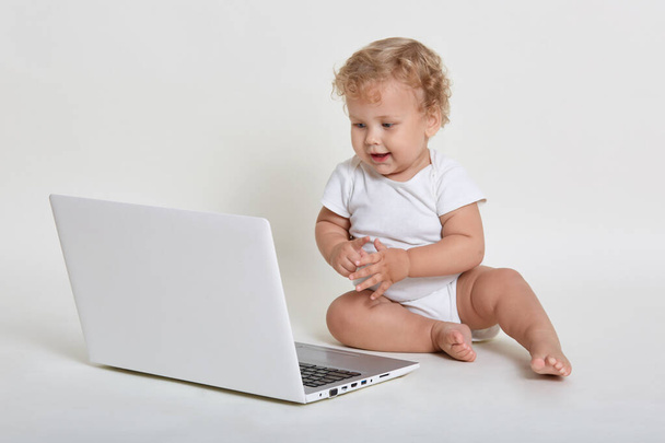 Little blond curly haired male wearing body suit, sitting on floor with lap top, looking at gadget screen with curious facial expression, posing isolated over white background. - Zdjęcie, obraz