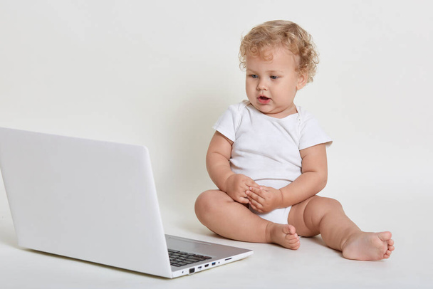 Little kid with laptop sitting on floor with opened mouth and looking directly at lap top screen, has concentrated look, dresses body suit, posing barefoot against white wall. - Valokuva, kuva