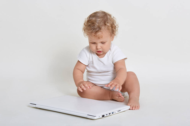 Little kid with laptop sitting on ground, closed laptop computer with great interest, curious infant plays barefoot with notebook isolated over white background. - Fotó, kép