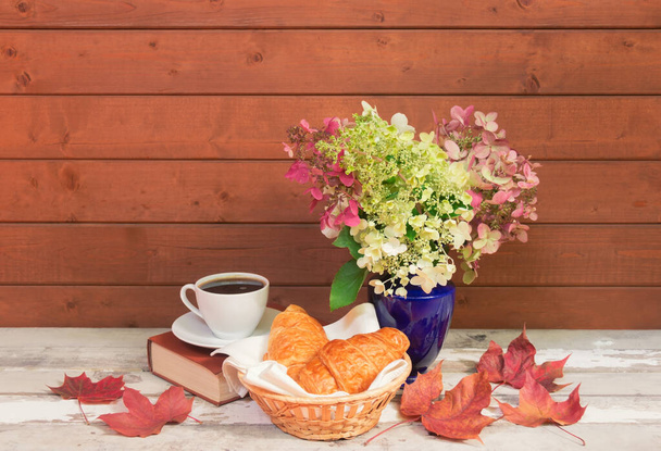 Xícara de café, croissants, livro e outonal desbotando flores de hortênsia rosa e branca em vaso azul na mesa de madeira envelhecida. Vista com espaço de cópia. - Foto, Imagem