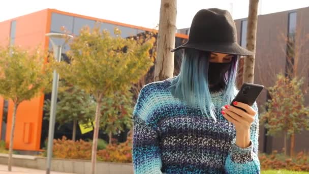 Студентка с голубыми волосами в защитной черной маске и шляпе со смартфоном. Городской модный костюм. COVID-19 - Кадры, видео