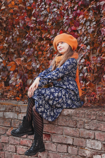 Beautiful girl in an orange beret sits putting one foot on the other. Beautiful girl on a background of red leaves - Photo, Image