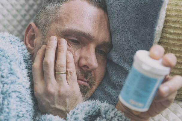 Mature man suffering from depression lies sadly covered with a blanket on his bed and holds a bottle of antidepressants in his hand - Photo, Image