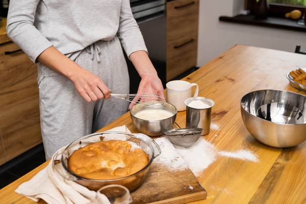 Tasty apple pie on a oaken wood table with a ingredients. Woman in a gray pajama cooking a delicious apple pie on a kitchen in a loft style. Milk, eggs, sugar and a baked pie on a table. Home kitchen. - Zdjęcie, obraz