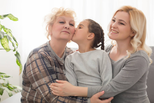 Three generations of women. Beautiful woman and teenage girl are kissing their granny while sitting on couch at home - Foto, Imagem