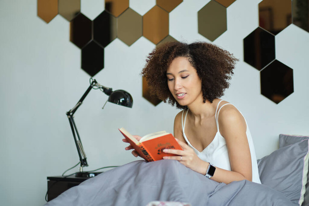 Νεαρή γυναίκα κρατά ένα βιβλίο στην κρεβατοκάμαρά της. - Φωτογραφία, εικόνα