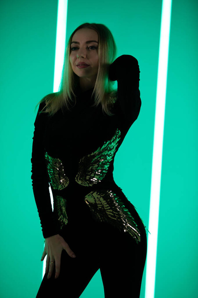 Blonde in a black tight-fitting suit with sequins poses against a background of neon lamps with green light. - Fotó, kép