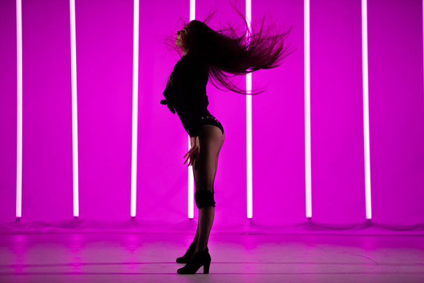 Side view of a young slender woman with flying hair posing standing in the studio against a background of neon lamps with purple backlight. - Photo, image