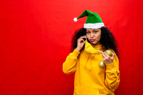 Hermosa mujer latina lleva la máscara protectora coronavirus, mientras usa un sombrero de Santa Claus y sostiene una esfera de árbol de Navidad. - Foto, imagen