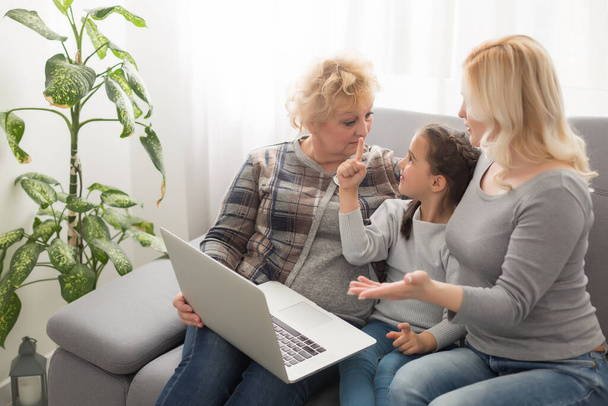 Happy three generations of women sit relax on couch laugh watching funny video on laptop, smiling positive females grandmother, mother and daughter have fun rest on sofa enjoy movie on computer - 写真・画像