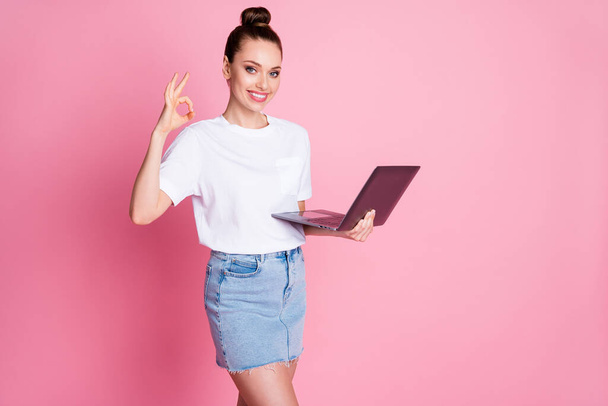 Photo of positive girl enjoy remote use laptop show okay sign wear white t-shirt denim jeans isolated over pastel color background - 写真・画像
