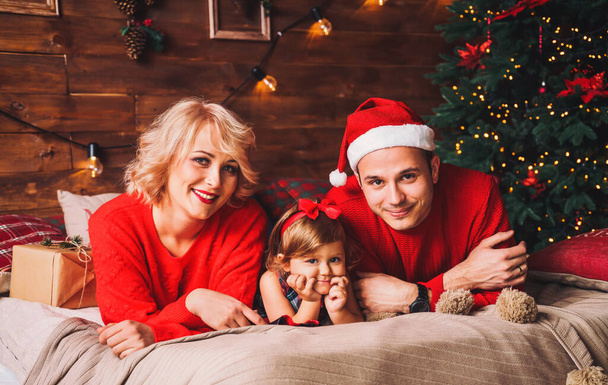 A család karácsonyi sapkában fekszik az ágyon. Anya, apa és a baba jól érzik magukat a nappaliban díszített karácsonyfa és ajándékdobozok. Szilveszter és karácsony ünnepek - Fotó, kép