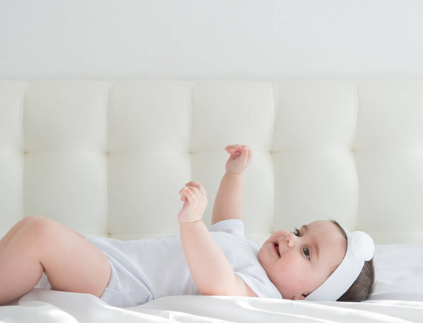 Nettes gesundes Mädchen 6 Monate lächelnd in einem weißen Body auf einem Bett auf weißem Bettzeug liegend. - Foto, Bild