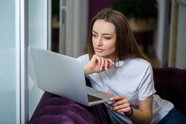 Uma jovem está sentada no sofá e trabalhando em um laptop remotamente do trabalho. Menina com um computador de joelhos olhando para o monitor - Foto, Imagem
