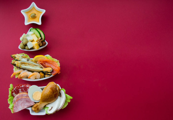 Bord in de vorm van een kerstboom met eiwit voedsel - vlees, vis, kazen, noten, enz. Rode achtergrond. Het concept van keto dieet traktaties voor de vakantie. Kopieerruimte. - Foto, afbeelding