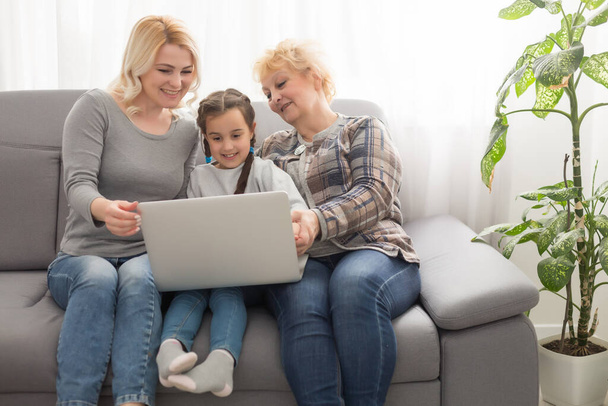 Happy three generations of women sit relax on couch laugh watching funny video on laptop, smiling positive females grandmother, mother and daughter have fun rest on sofa enjoy movie on computer - Zdjęcie, obraz
