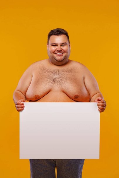 Fat and happy shirtless man displaying a white banner ad isolated over a yellow background. - Фото, изображение
