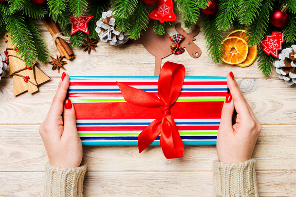 Κάτοψη μιας γυναίκας που κρατάει ένα κουτί δώρου στα χέρια της σε εορταστικό ξύλινο φόντο. Δέντρα και χριστουγεννιάτικα στολίδια. Έννοια του νέου έτους. - Φωτογραφία, εικόνα