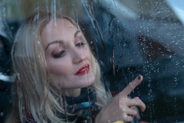 looks through the wet glass, drops a girl from the car window on the rain, with a Caucasian appearance in a black car. smiling at camera - Φωτογραφία, εικόνα