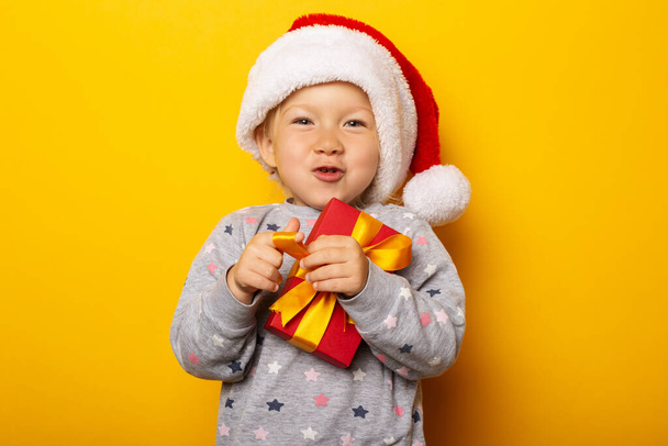 Dítě v klobouku Santa Clause má dar na žlutém pozadí. Dárek k Vánocům, vánoční ráno. Nápis. - Fotografie, Obrázek