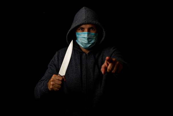Νεαρός άνδρας με ιατρική μάσκα δείχνει το δάχτυλο ενώ κρατάει μαχαίρι σε μαύρο φόντο - Φωτογραφία, εικόνα
