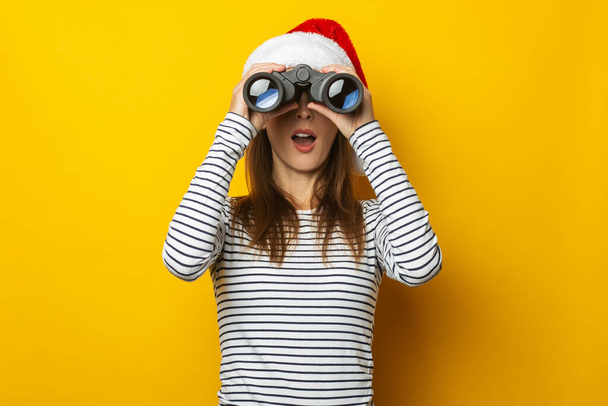 Молода жінка з здивованим обличчям у капелюсі Санта-Клауса дивиться через бінокль на жовтому тлі. Банер. - Фото, зображення
