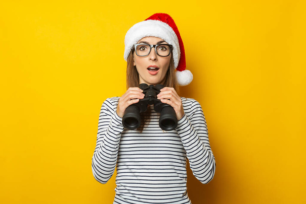 Молодая женщина с удивленным лицом в шляпе Санта-Клауса держит бинокль на желтом фоне. Баннер. - Фото, изображение