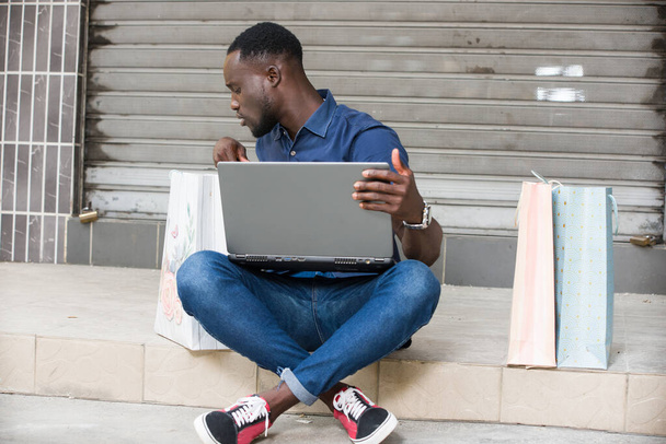 jeune homme assis sur le sol avec ordinateur portable regarder dans le sac à provisions. - Photo, image